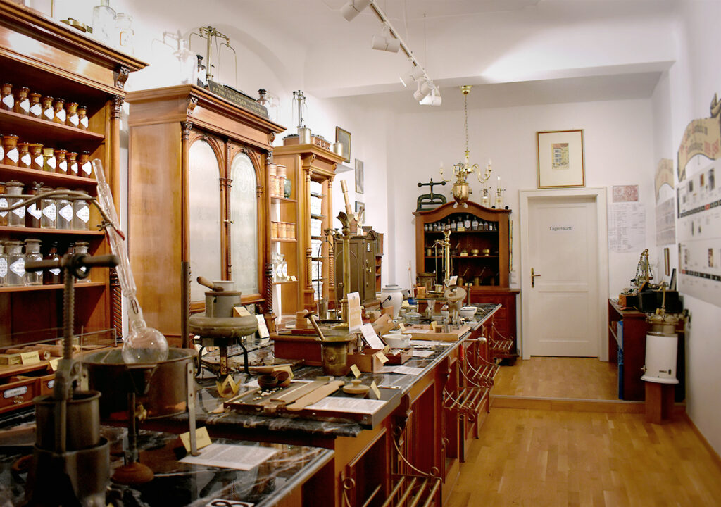 Der Hauptraum des Adler Apotheke Museums zeigt eine historische Apotheke mit Holzregalen, die mit verschiedenen Gefäßen und alten pharmazeutischen Utensilien gefüllt sind. In der Mitte des Raumes stehen Holztische, auf denen zahlreiche Objekte ausgestellt sind. An den Wänden hängen Bilder und Dokumente, die die Geschichte der Apotheke veranschaulichen.