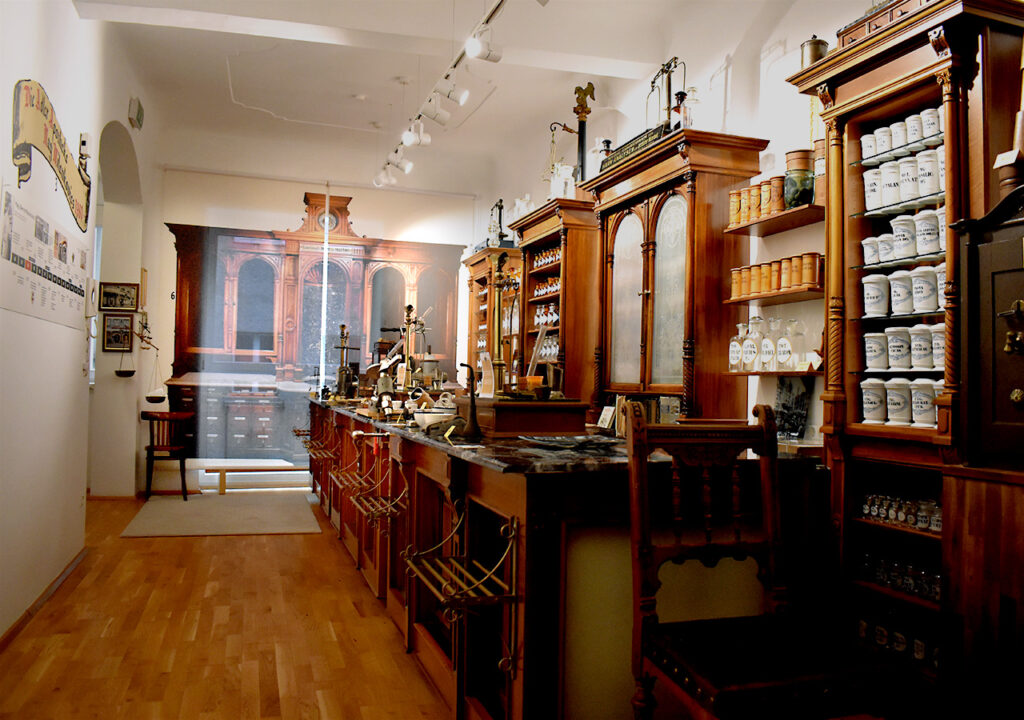 Der Hauptraum des Adler Apotheke Museums zeigt eine historische Apotheke. In der Mitte des Raumes stehen Holztische, auf denen zahlreiche Objekte ausgestellt sind. An der linken Wand hängt ein Zeitstrahl, der die Geschichte der Apotheke veranschaulicht.