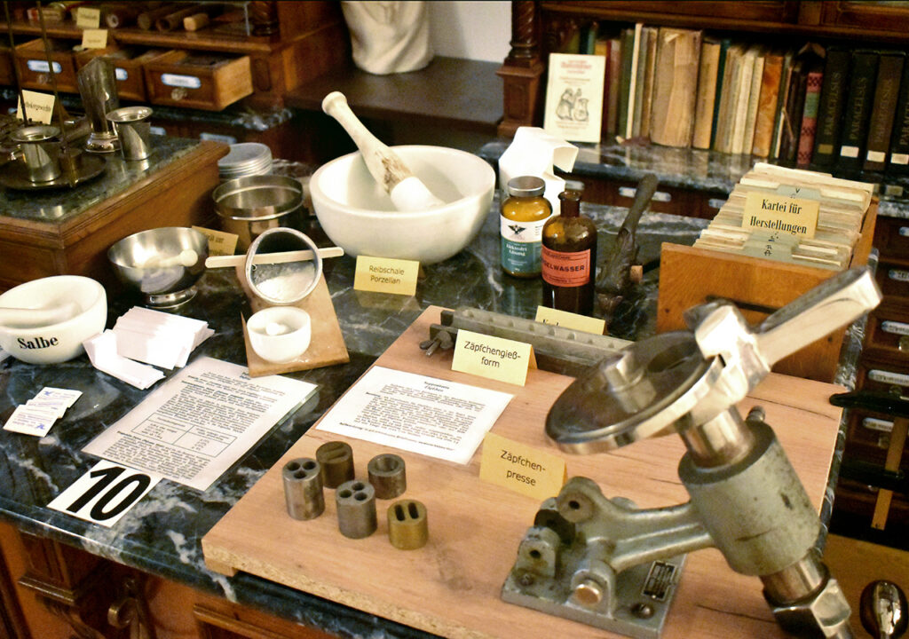 Eine Nahaufnahme von verschiedenen pharmazeutischen Utensilien im Adler Apotheke Museum. Auf einem Marmor-Tisch stehen Schalen, ein Mörser, sowie verschiedene Flaschen mit Etiketten. Diverse Informationsblätter sind ebenfalls ausgelegt, die die Verwendung der Objekte erklären