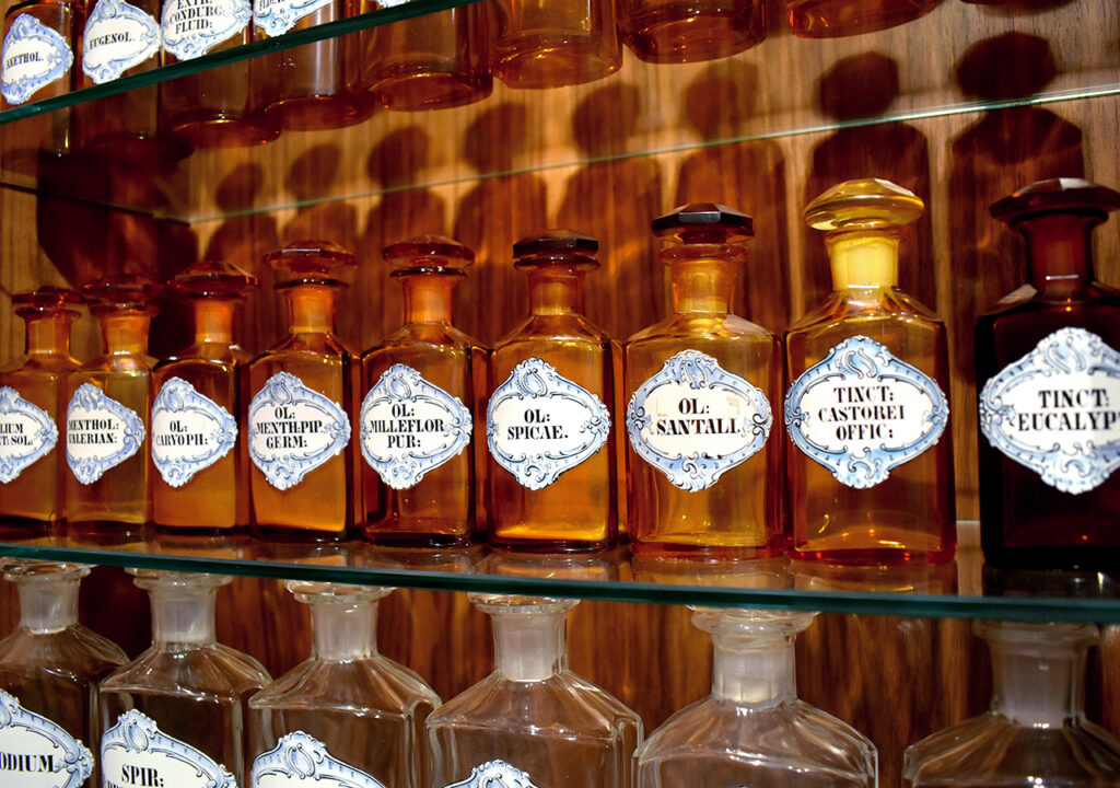 Eine Reihe von historischen Apothekerflaschen im Adler Apotheke Museum. Die Flaschen sind in verschiedenen Formen und Größen. Jede Flasche ist mit einem eleganten Etikett versehen, das den Inhalt beschreibt, darunter Begriffe wie „Öl Spicae“ und „Tinktur Eucalyptus“. Die Flaschen stehen auf einem Regal, das interessante Schatten wirft.