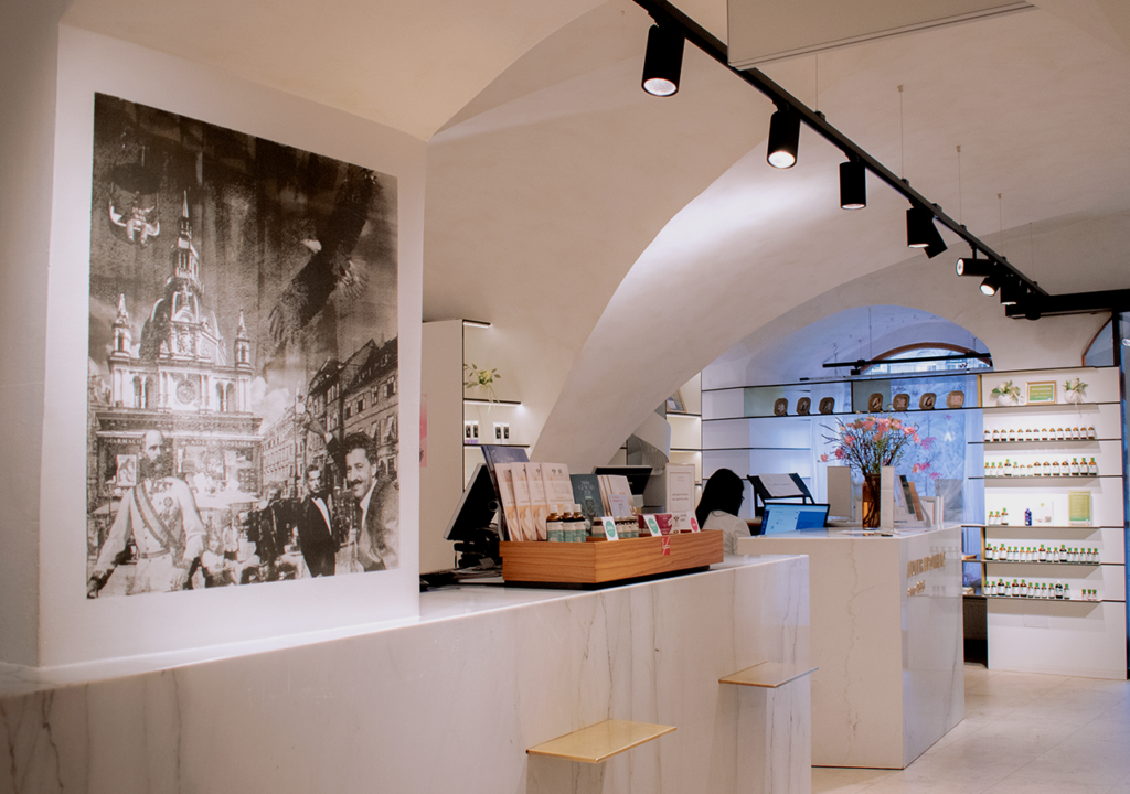 Blick ins Innere der Adler Apotheke, das eine elegante und moderne Gestaltung zeigt. An der Wand befindet sich ein großes Schwarz-Weiß-Bild, das eine historische Szenerie darstellt. Der Empfangstresen aus marmoriertem Material ist gut sichtbar, während im Hintergrund Regale mit verschiedenen Produkten angeordnet sind. Sanftes Licht von den Deckenleuchten schafft eine einladende Atmosphäre.