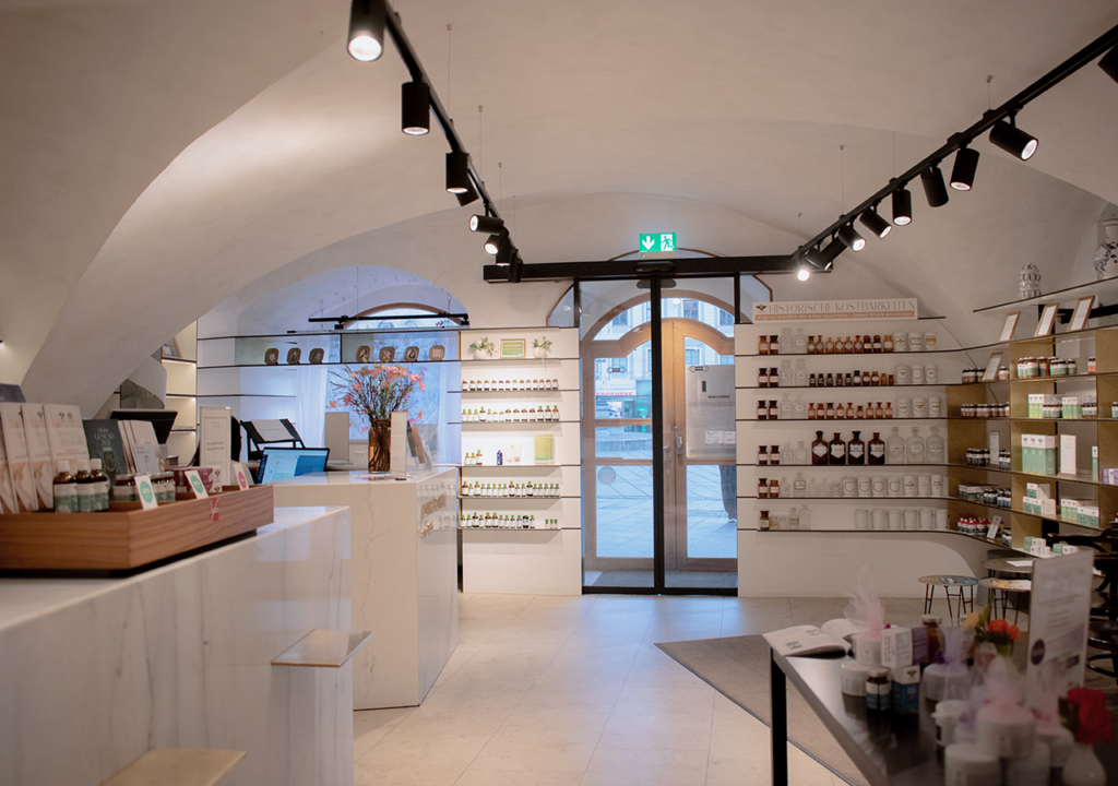 Blick in das Innere der Adler Apotheke mit einem modernen und einladenden Ambiente. Der Tresen aus Marmor steht im Vordergrund, während an den Wänden Regale mit verschiedenen Produkten sichtbar sind. Durch die große Glastür im Hintergrund strömt Licht herein, und im Raum befinden sich einige dekorative Pflanzen sowie eine angenehme Anordnung von Waren.