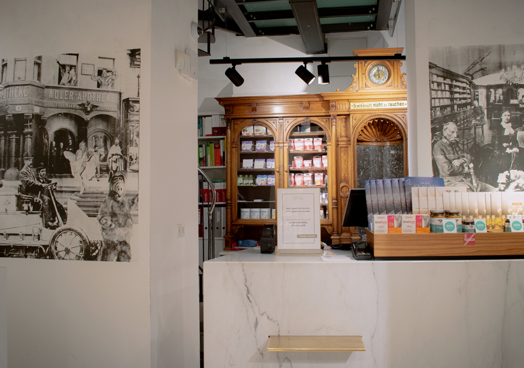 Der Empfangsbereich der Adler Apotheke mit einer modernen Theke aus Marmor. Im Hintergrund ist ein historisches Holzregal mit einer Uhr und vielen Packungen sichtbar. An der Wand sind Schwarzweiß-Fotos von historischen Apothekenkunden sowie einer alten Apotheke angebracht. Der Raum strahlt eine Kombination aus klassischem und modernem Design aus.