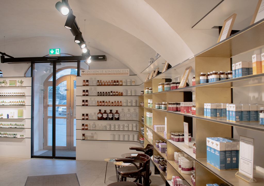 Innenansicht der Adler Apotheke mit hellen Regalen, die verschiedenen Pflegeprodukten und Medikamenten ausgestellt sind. An der linken Wand sind zahlreiche Flaschen und Gläser auf Regalen angeordnet, während die rechte Wand leerere Regale für zusätzliche Produkte bietet. Der Eingangsbereich ist durch eine große Glastür sichtbar, die viel Tageslicht hereinzulassen. Stühle stehen bereit, um den Kunden einen komfortablen Platz zu bieten.