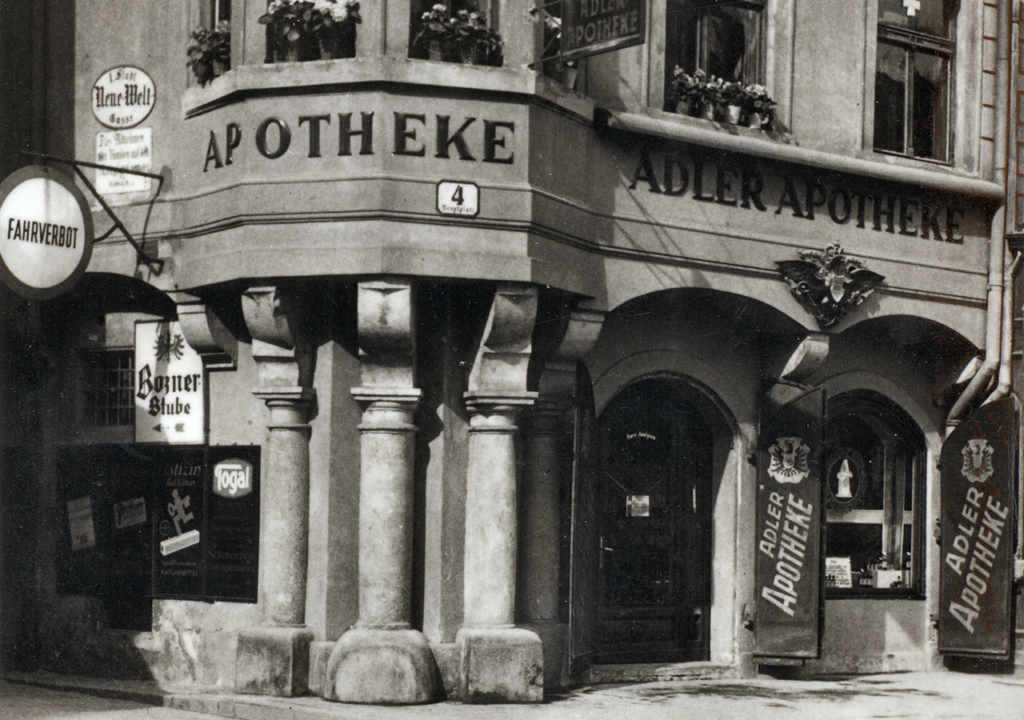 Historisches Schwarz-Weiß-Foto der Adler Apotheke aus den 1930er Jahren. Die markante Fassade zeigt das Schild "ADLER APOTHEKE" sowie weitere Werbeschilder. Die Architektur ist im typischen Stil der Zeit gehalten, mit dekorativen Elementen und Säulen. Die Straßenansicht vermittelt einen nostalgischen Eindruck des städtischen Lebens in dieser Epoche.