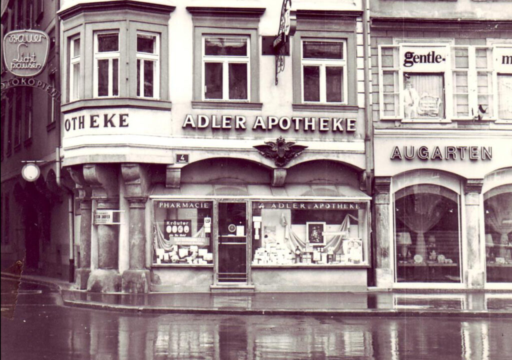 Historisches Schwarz-Weiß-Foto der Adler Apotheke aus den 1950er Jahren. Die Fassade zeigt das Schild "ADLER APOTHEKE" und ein Schaufenster mit verschiedenen Produkten, die zum Verkauf angeboten werden. Die Architektur spiegelt den Stil der Nachkriegszeit wider, und die nassen Straßen deuten auf einen Regen hin, der der Szene einen nostalgischen Charakter verleiht.