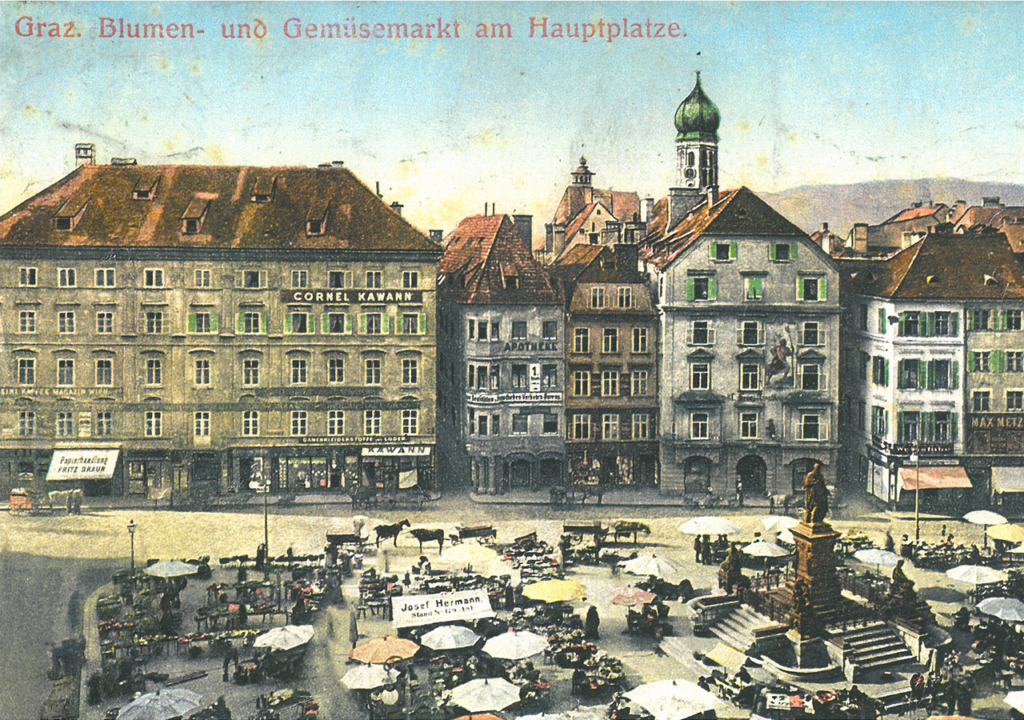 Historische Postkartenansicht des Grazer Hauptplatzes, auf der die Adler Apotheke gut sichtbar ist. Die lebendige Szene zeigt den Blumen- und Gemüsemarkt mit Ständen, Tischen und vielen Menschen. Umgeben von historischen Gebäuden, darunter die Apotheke, vermittelt das Bild einen Eindruck vom städtischen Leben und der Architektur in Graz zu dieser Zeit.