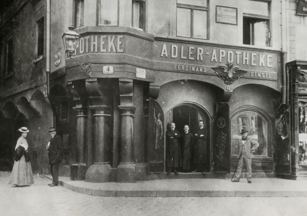 Historisches Schwarz-Weiß-Foto der Adler Apotheke um 1900. Die Fassade der Apotheke mit dem Schild "ADLER-APOTHEKE" ist prominent zu sehen. Vor dem Eingang stehen drei Männer in historischen Gewändern, während eine Frau in einem eleganten Kleid auf der Straße steht. Die Szene vermittelt einen Eindruck des städtischen Lebens und der Architektur der damaligen Zeit.