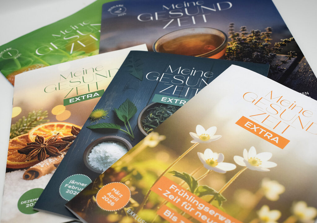 Eine Nahaufnahme von mehreren Ausgaben des Extrahefts mit Aktionen und der Zeitschrift "Meine Gesundzeit" von der Adler Apotheke für Vorteils-Kunden. Die Zeitschriften zeigen frische grüne Pflanzen und eine Tasse Tee.