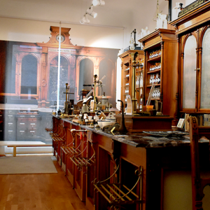 Das Bild zeigt den Hauptraum des Adler Apotheken Museums. Historische Artefakte der Adler Apotheke sind am rechten Seitenrand zu sehen, wo ein Holztresen steht, der mit verschiedenen pharmazeutischen Werkzeugen und Glasgefäßen ausgestattet ist. Auf der linken Seite kann man eine Zeitstrahl sehen.