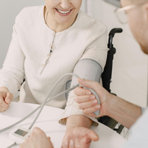 Eine Person lächelt, während ihr Blutdruck von einem Gesundheitsdienstleister gemessen wird. Der Dienstleister hält das Blutdruckmessgerät in der Hand, während die Patientin entspannt am Tisch sitzt.