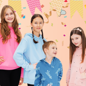 Vier Kinder stehen lächelnd vor einem bunten, festlichen Geburtshintergrund mit Konfetti und bunten Wimpeln. Zwei Mädchen tragen pinke und hellblaue Hoodies, während ein Junge in einem blauen Hoodie und ein weiteres Mädchen in einem rosa Hoodie gekleidet ist.