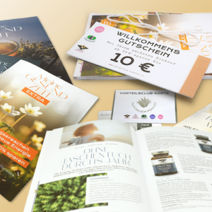 Gestreute Drucksachen auf hellem Untergrund: mehrere Ausgaben der „Adler Apotheke“ Zeitungen, ein Extraheft, ein aufgelegter Haus­spezialitäten‑Katalog, eine Vorteilsclub‑Karte und ein Willkommenspaket mit 10 € Gutschein