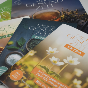 Eine Nahaufnahme von mehreren Ausgaben des Extrahefts mit Aktionen und der Zeitschrift "Meine Gesundzeit" von der Adler Apotheke für Vorteils-Kunden. Die Zeitschriften zeigen frische grüne Pflanzen und eine Tasse Tee.