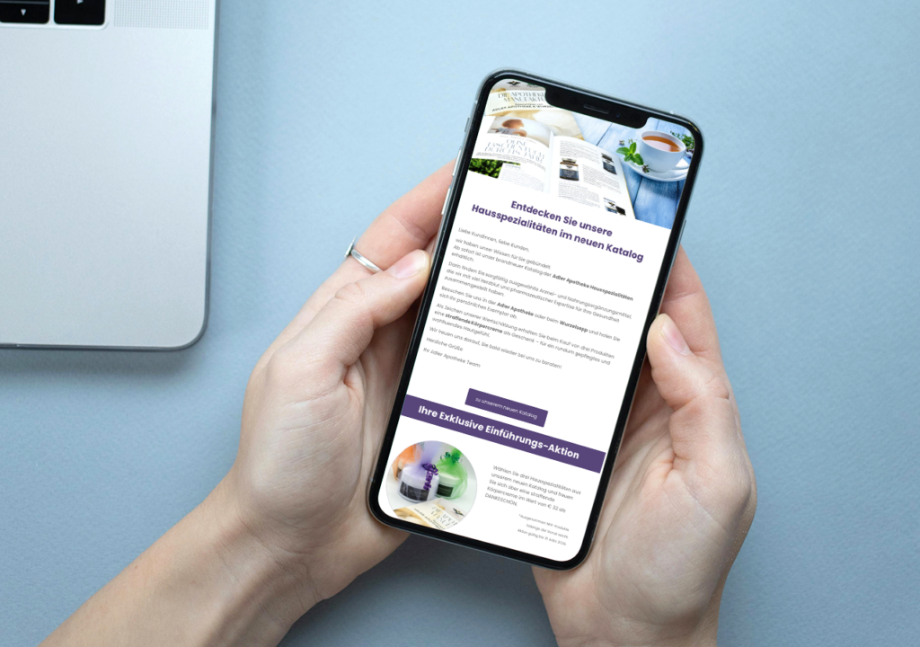 Hände halten ein Smartphone über einem hellblauen Tisch; auf dem Bildschirm ist der Newsletter der Adler Apotheke mit Textblöcken, Bildern von Produkten und einem lila Call-to-Action-Button zu sehen.