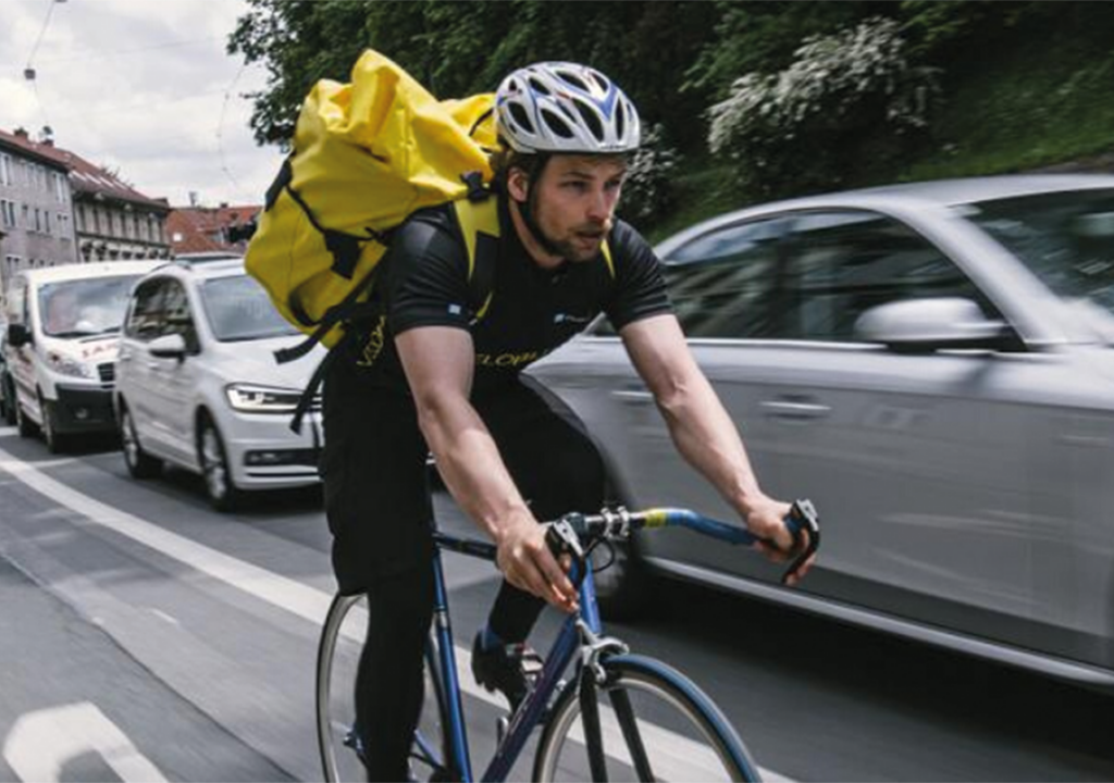 Ein Radbote hat einen gelben Rucksack am Rücken und fährt gerade durch die Grazer Innenstadt.