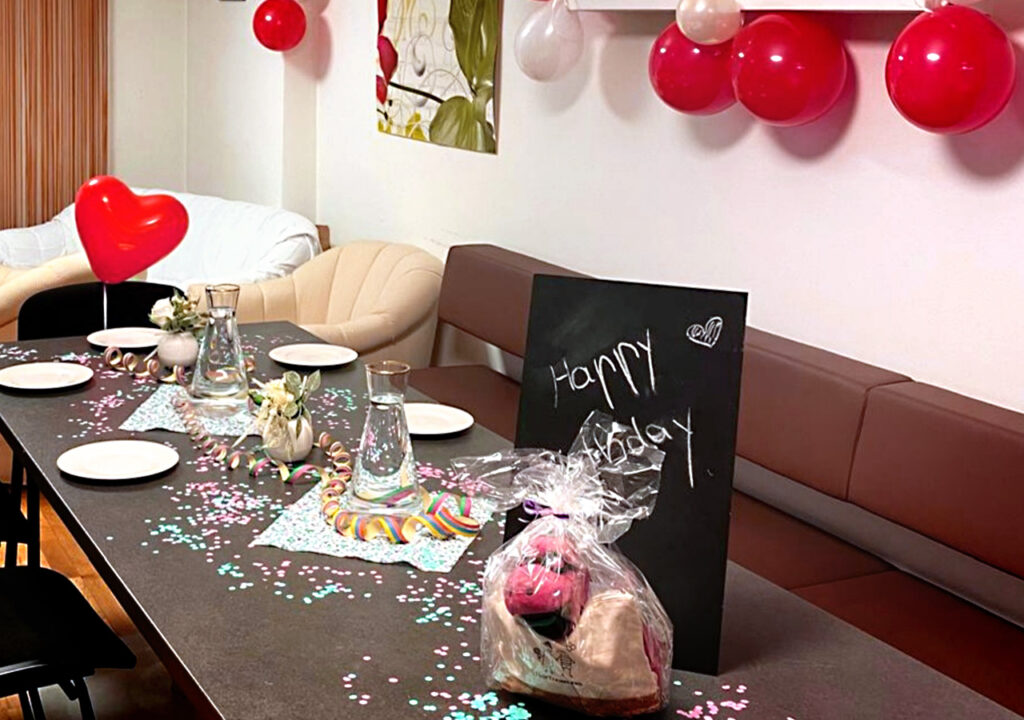 Festlich gedeckter Tisch in der Adler‑Apotheke mit Konfetti, Girlanden und roten Luftballons; auf dem Tisch Teller, Gläser, eine Herz‑Luftballon‑Dekoration, ein als Geschenk verpacktes Stofftier und eine schwarze Kreidetafel mit der Aufschrift „Happy Birthday“.