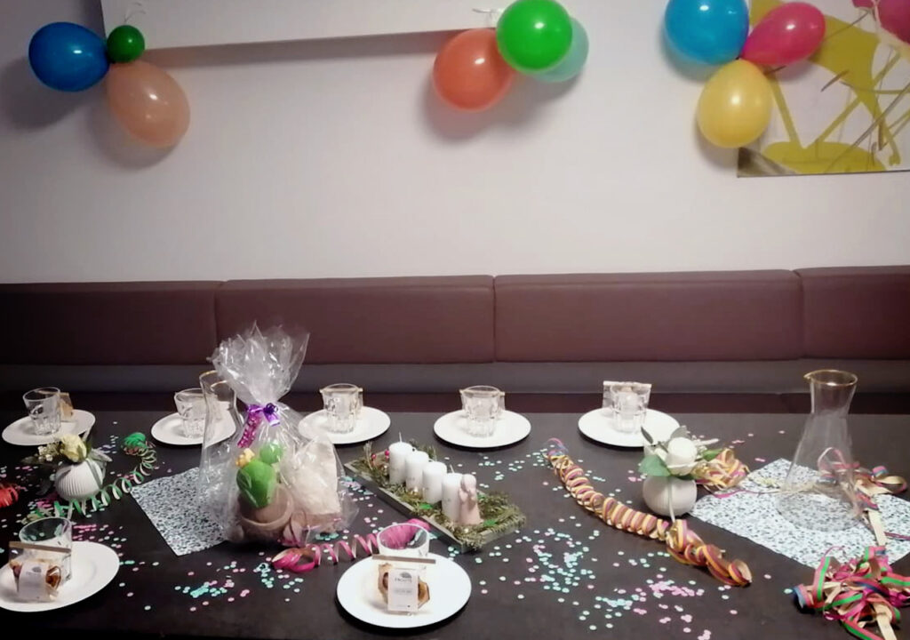 Ein festlich dekorierter Tisch in einer Apotheke: Auf einem dunklen Tisch sind mehrere Gedecke mit kleinen Desserttellern und Glasstampern arrangiert; Konfetti, bunte Luftschlangen und Geschenkpakete liegen verstreut. In der Mitte stehen verschenkte Präsente (z. B. ein in Zellophan eingepacktes Geschenk, kleine Kerzen) und dekorative Blüten. Im Hintergrund eine braune Polsterbank und bunte Luftballons, die an der Wand hängen.