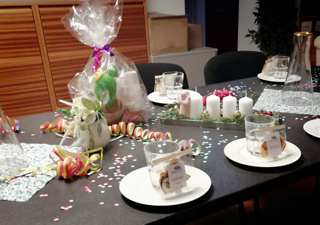 Festlich gedeckter Tisch in der Adler‑Apotheke mit Konfetti und Girlanden; auf dem Tisch kleine Teller mit einzeln verpackten Gebäckstücken, Gläser, Karaffen, ein verpacktes Geschenk, Kerzenarrangement und Blumen in Vasen.