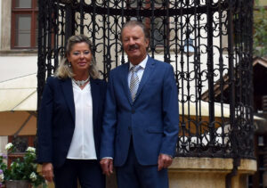 Silvia Milenkovics und Bernd Milenkovics stehen lächelnd in eleganter Kleidung nebeneinander im historischen Landhaushof in Graz vor dem kunstvollen, geschmiedeten Brunnen. Bernd Milenkovics trägt einen blauen Anzug mit gestreifter Krawatte, Silvia Milenkovics einen dunklen Hosenanzug mit weißer Bluse.