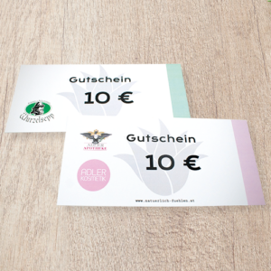Eine Nahaufnahme von zwei physischen Geschenkgutscheinen der Adler Apotheke, präsentiert im quadratischen Format als Icon_Gutschein_500x500.png. Die Gutscheine liegen auf einem hellbraunen Holztisch. Der vordere Gutschein ist pink und weiß, der hintere mintgrün und weiß. Beide zeigen deutlich den Text „Gutschein“ und den Wert „10 €“ in schwarzer Schrift. Der vordere Gutschein trägt das pinkfarbene kreisförmige Logo „Adler Kosmetik“ und den Hinweis „Exklusiv für Vorteilscardmitglieder“. Die Gutscheine haben ein fächerartiges florales Design im Hintergrund
