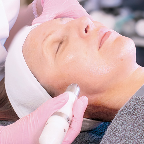 Eine Kosmetikerin in weißer Arbeitskleidung behandelt eine liegende Kundin im Adler Kosmetikstudio mit Dermadrop, dabei sind Handschuhe und Schutzmaske zu sehen, im Hintergrund Deckenlampen und Geräte auf dem Behandlungstisch.