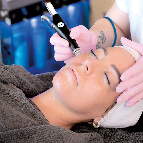 Eine Kosmetikerin behandelt eine Kundin im Adler Kosmetikstudio mit einer Hydrafacial-Behandlung: Die Kundin liegt entspannt auf dem Behandlungstisch, während die Kosmetikerin mit Handschuhen das Gerät an der Haut anlegt; im Hintergrund sind die Hydrafacial-Geräte und Flüssigkeitsaufsätze zu sehen.