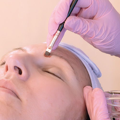Professionelle Augenbrauen- und Wimpernbehandlung im Kosmetikstudio: Eine entspannte Kundin mit weißem Stirnband erhält eine präzise Augenbrauen-Styling-Behandlung. Die Kosmetikerin trägt rosa Handschuhe und trägt mit einem abgeschrägten Kosmetikpinsel feine Farbe auf die Braue auf, während sie in der anderen Hand ein kleines Glasschälchen hält.